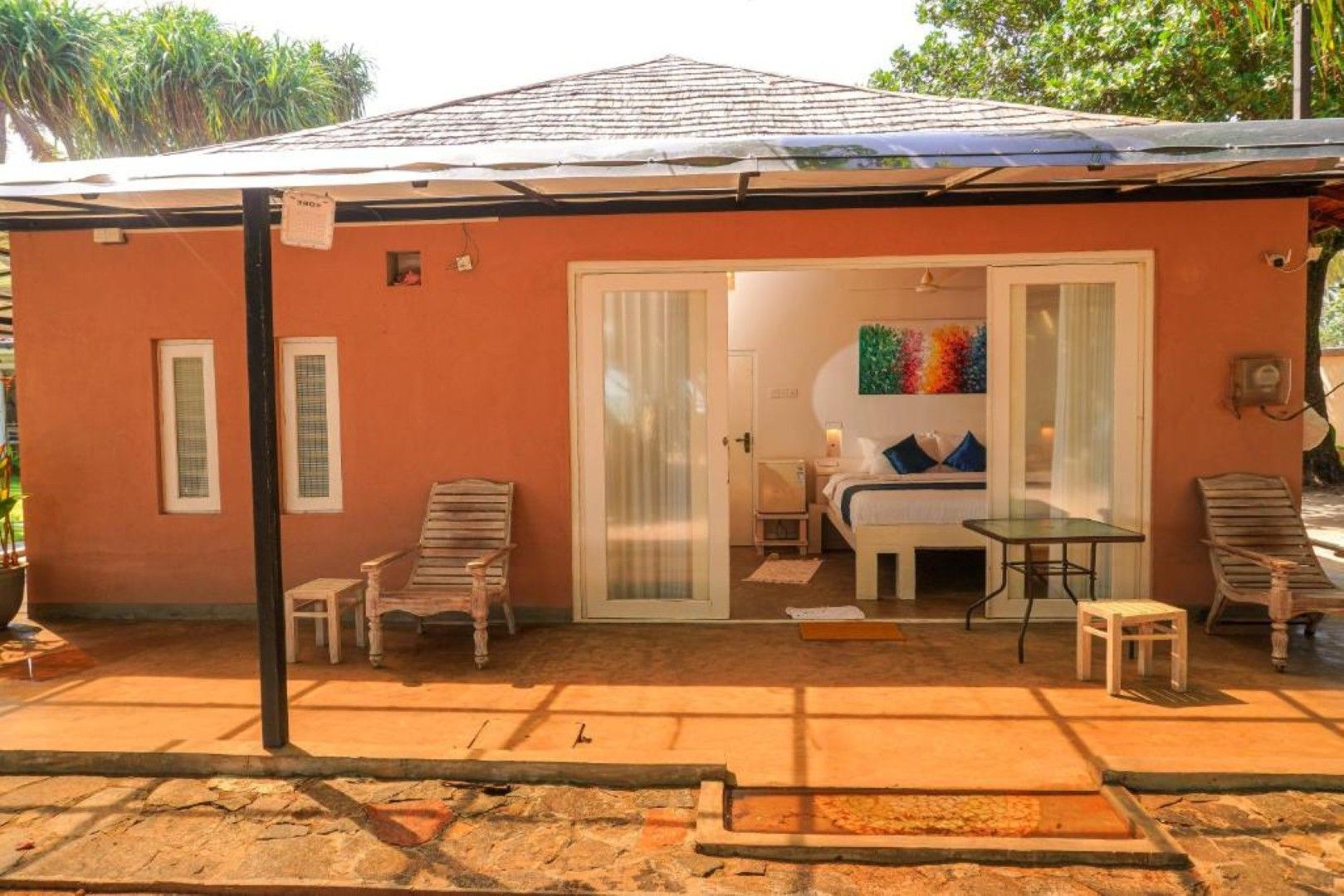 A small, single-story building with a tiled roof, a covered patio area with a table and chairs, and a sliding glass door leading to a bedroom with a bed and a colorful painting.