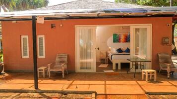A small, single-story building with a tiled roof, a covered patio area with a table and chairs, and a sliding glass door leading to a bedroom with a bed and a colorful painting.