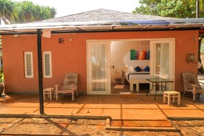 A small, single-story building with a tiled roof, a covered patio area with a table and chairs, and a sliding glass door leading to a bedroom with a bed and a colorful painting.