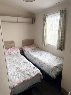 Une chambre de dortoir avec deux lits simples, une fenêtre avec des rideaux à motifs et une bouche d’aération au plafond.