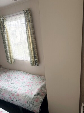 Ein Einzelbett mit Blumenmusterdecke, ein Fenster mit gemusterten Vorhängen und eine schlichte Wand.