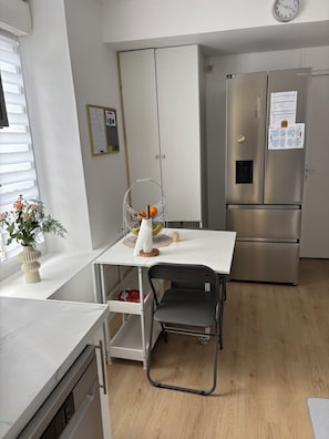 A modern kitchen with a stainless steel refrigerator, a white dining table with a chair, and a potted plant on the counter. - L'Original Duplex - Centre Ville (Liancourt)