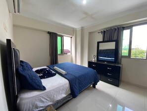 Ein Schlafzimmer mit Bett, Fernseher, Kommode und einem Fenster mit Vorhängen.