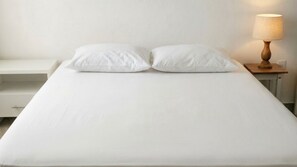 Ein ordentlich bezogenes Bett mit weißer Bettwäsche und Kissen, ein Nachttisch mit Lampe sowie eine Kommode zur Linken.