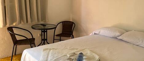 A hotel room with a bed, a small table, two chairs, an air conditioning unit, and a window with curtains.