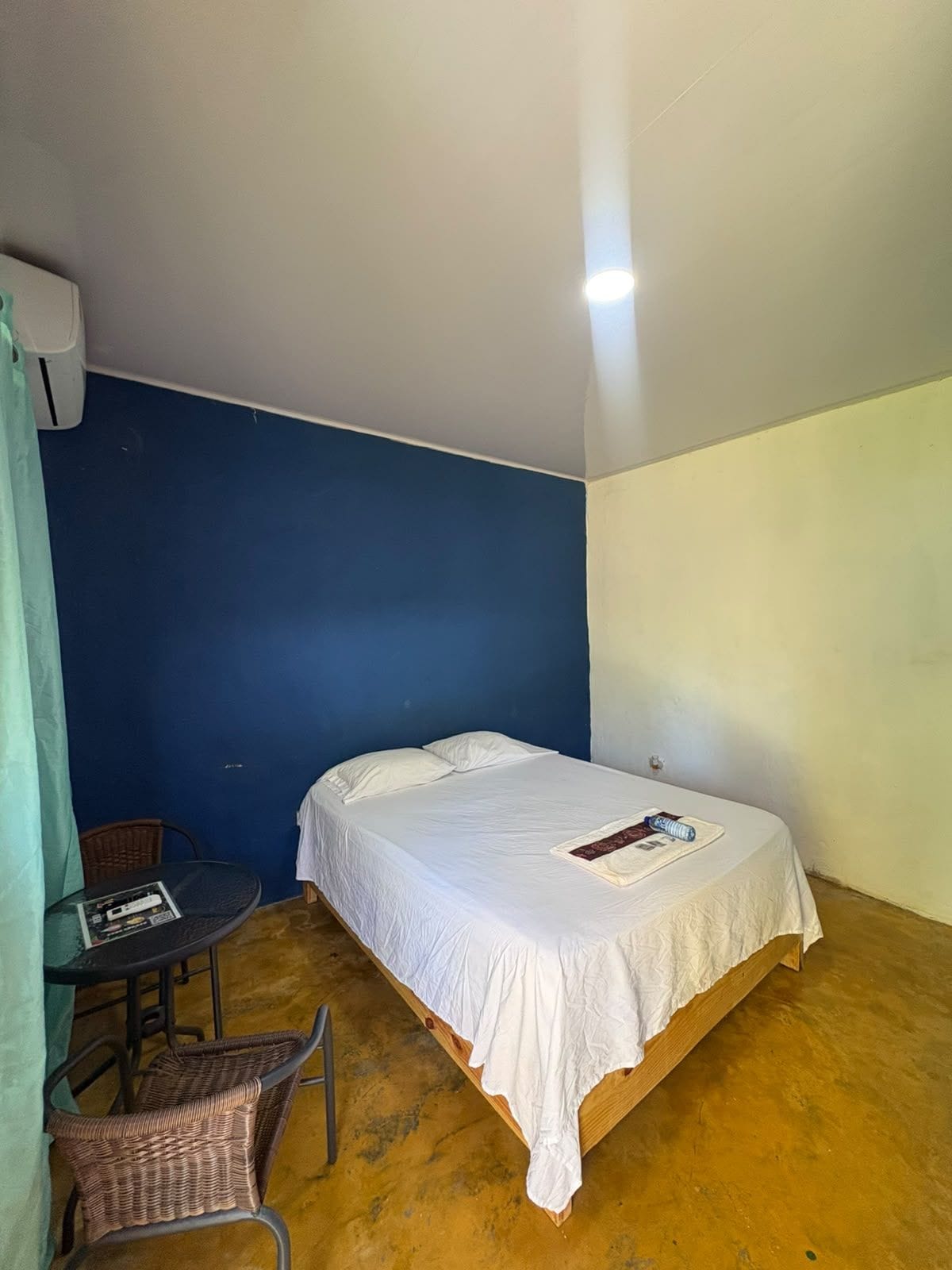 Een eenpersoonsbed met witte lakens, een klein tafeltje met een tijdschrift en een rieten stoel in een kamer met blauwe en witte wanden.