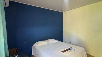 Een eenpersoonsbed met witte lakens, een klein tafeltje met een tijdschrift en een rieten stoel in een kamer met blauwe en witte wanden.