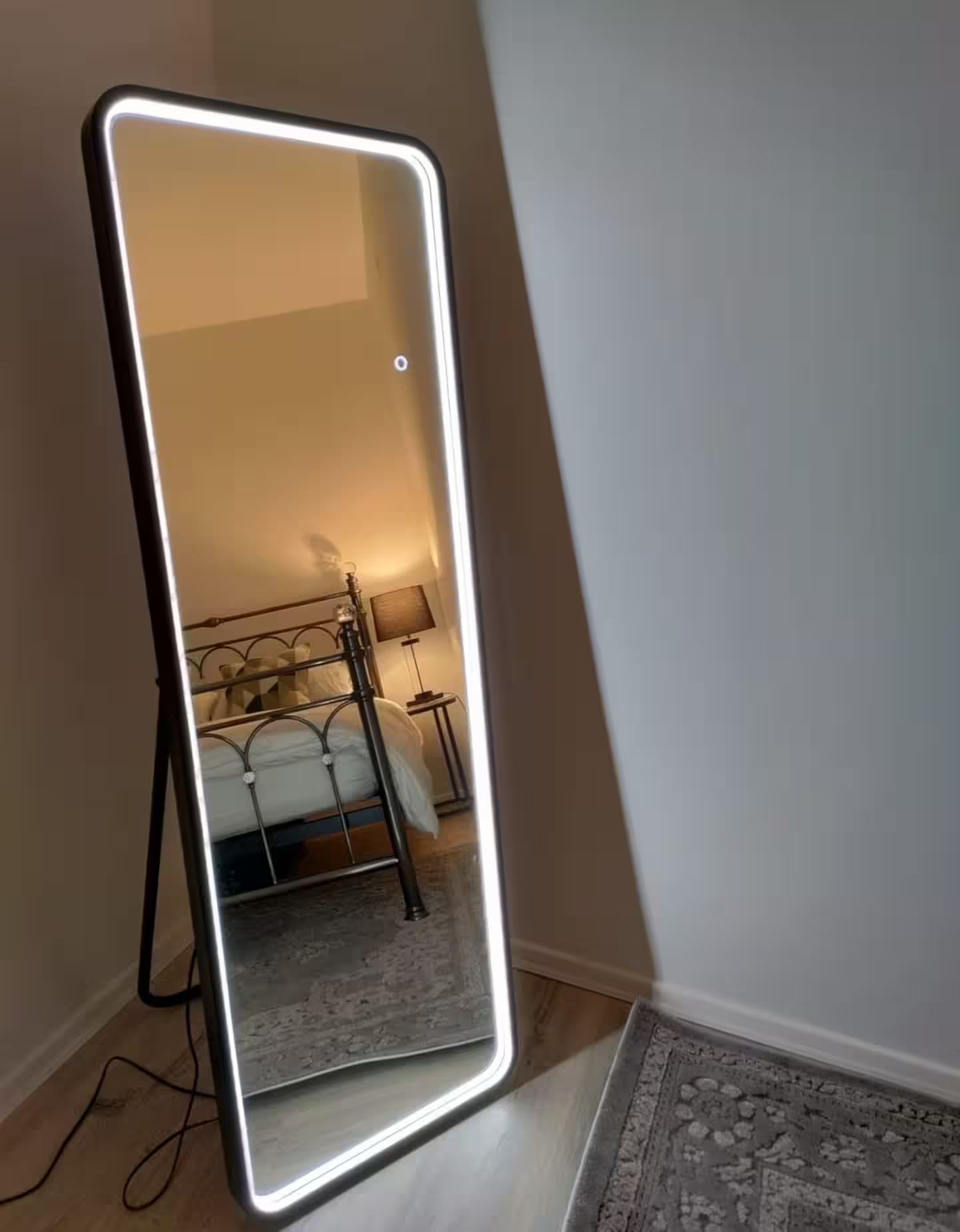 A full-length mirror with a black frame and LED lighting, reflecting a bed and a bedside lamp in a room with a wooden floor.