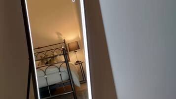 A full-length mirror with a black frame and LED lighting, reflecting a bed and a bedside lamp in a room with a wooden floor.