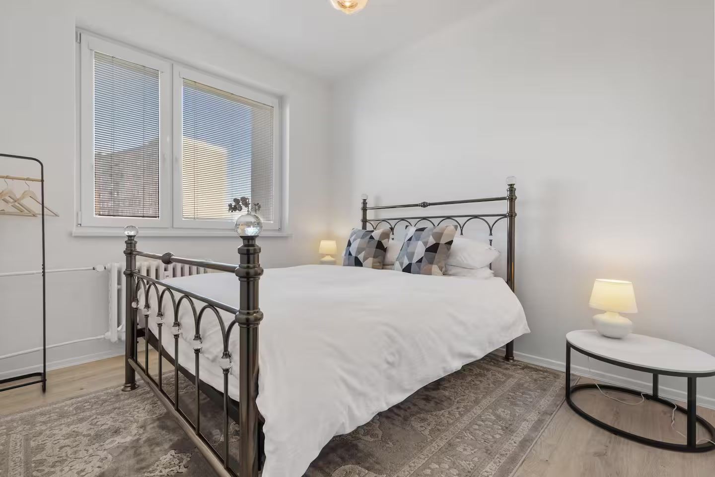 Een slaapkamer met een bed met metalen frame, witte beddengoed, een rond nachtkastje met een lamp en een raam met zonwering.