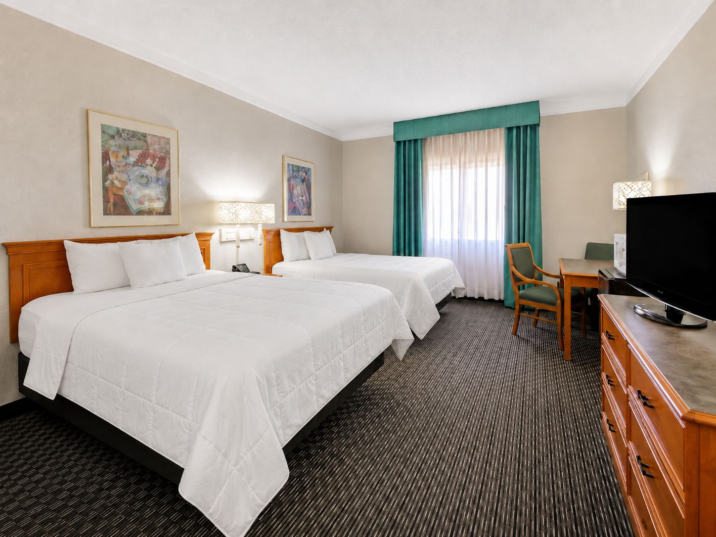 Quarto de hotel com duas camas, uma escrivaninha, televisão e obras de arte nas paredes.