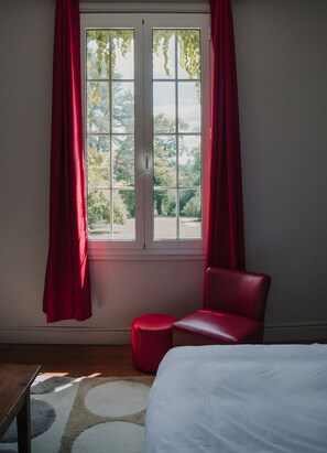 Ein Schlafzimmer mit einem Bett, einem roten Sessel, einem kleinen Hocker und einem Fenster mit Vorhängen.
