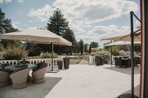 Een terras buiten met rieten meubilair, twee grote parasols en uitzicht op een tuin met bomen en een vijver.