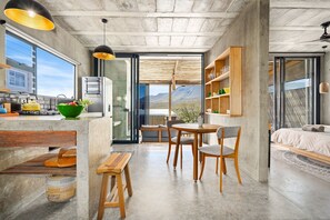 A modern kitchen with a large window, a dining area with a wooden table and chairs, and a bedroom with a bed and a view of the mountains. - KOH Pringle Pod (Pringle Bay)