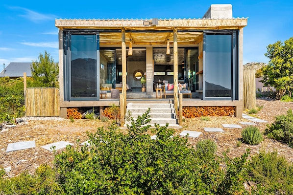 A modern house with large glass windows, a wooden deck, and a stone pathway leading to the entrance. - KOH Pringle Pod (Pringle Bay)