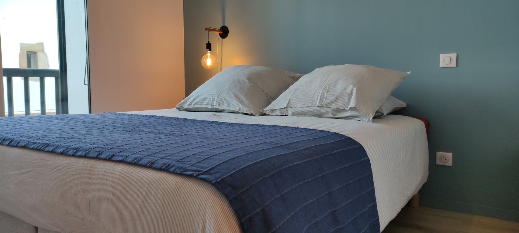 Ein Bett mit blauer Decke, zwei weißen Kissen und einer Wandleuchte.