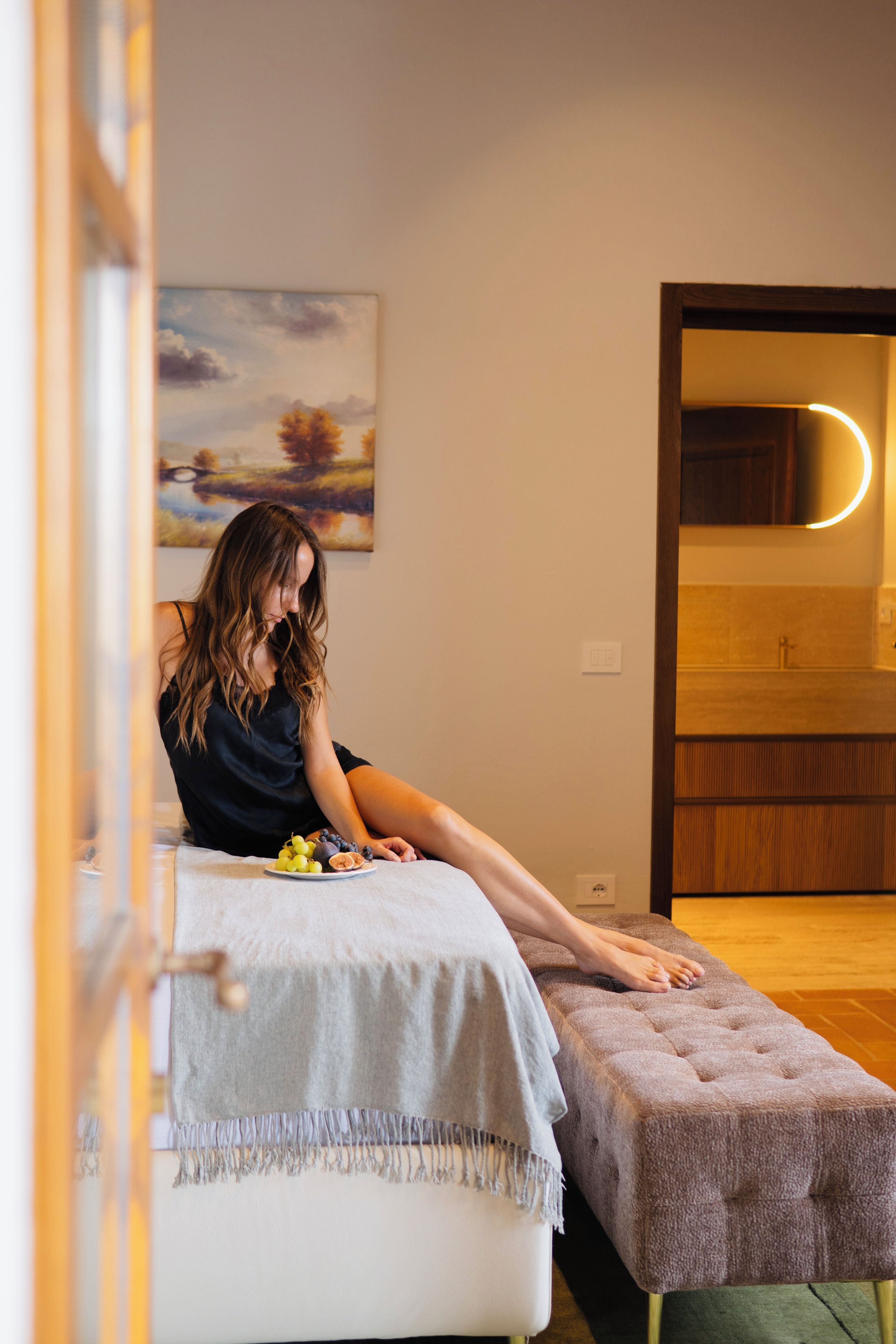 A person sitting on a bed with a plate of fruit, a painting on the wall, and a bathroom visible through an open door.