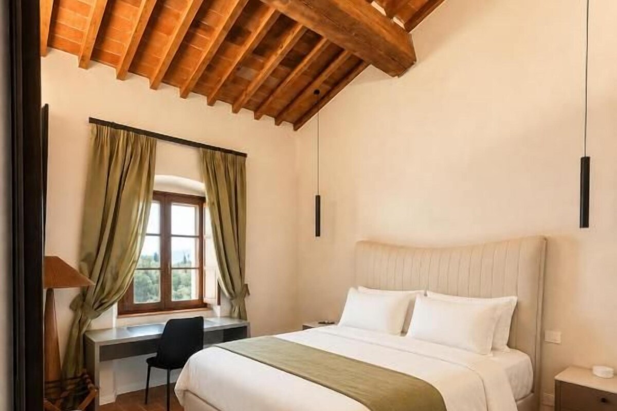 A bedroom with a large bed, a desk, and a chair. There is a window with curtains, a bench, and a hanging light fixture.