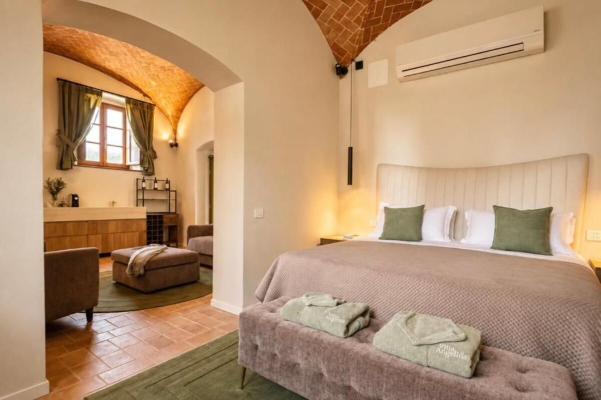 A hotel room with a bed, a sitting area with a chair and ottoman, a small kitchenette, and a brick archway.