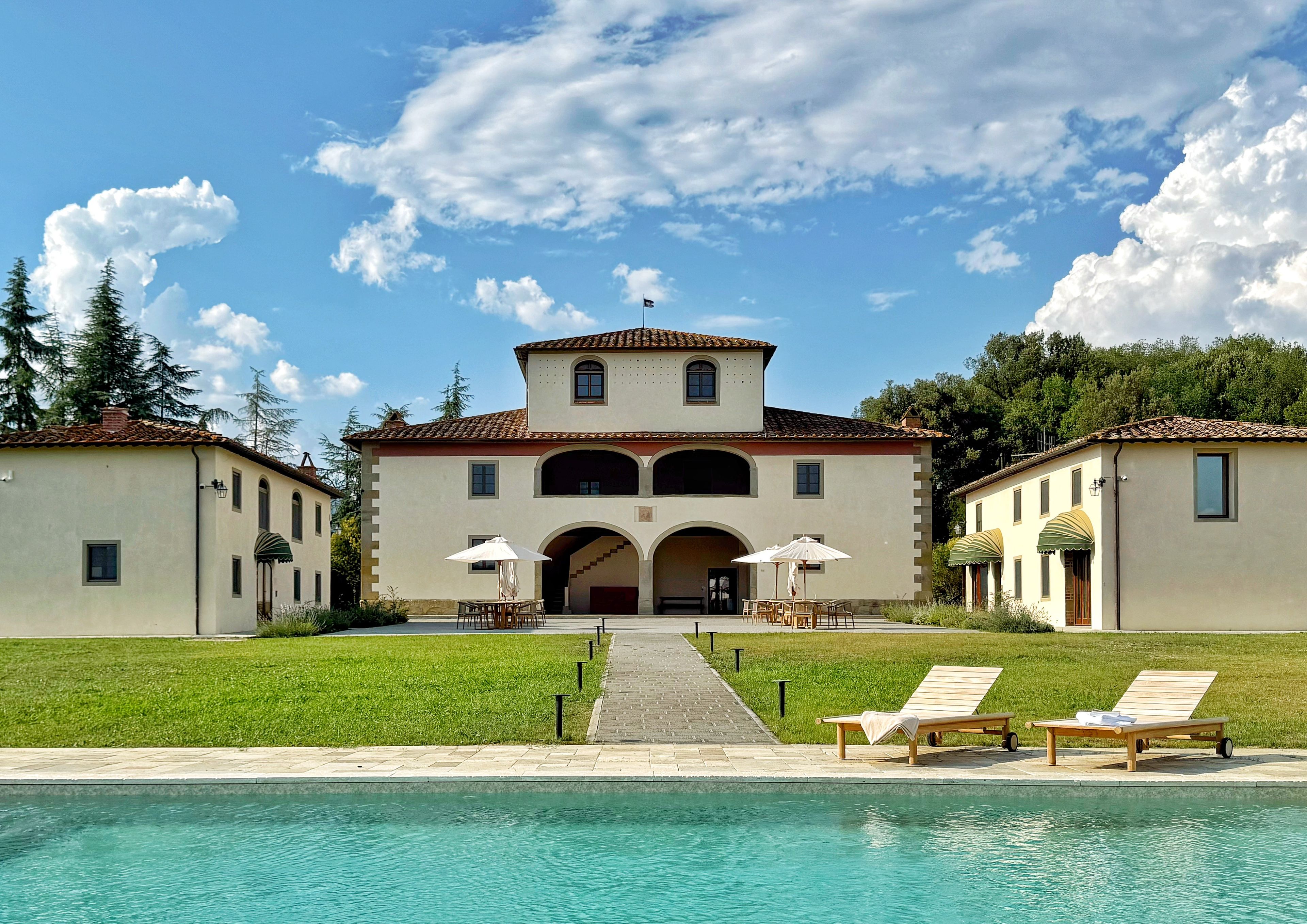 A two-story building with a swimming pool in front, featuring a covered patio area with chairs and umbrellas.