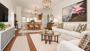 A spacious living room with a glass coffee table, a white sofa, and a dining area with a wooden table and chairs. - SHELL HOUSE - Old Florida Charm - Minutes to Historic Downtown Stuart & Beaches (Stuart)