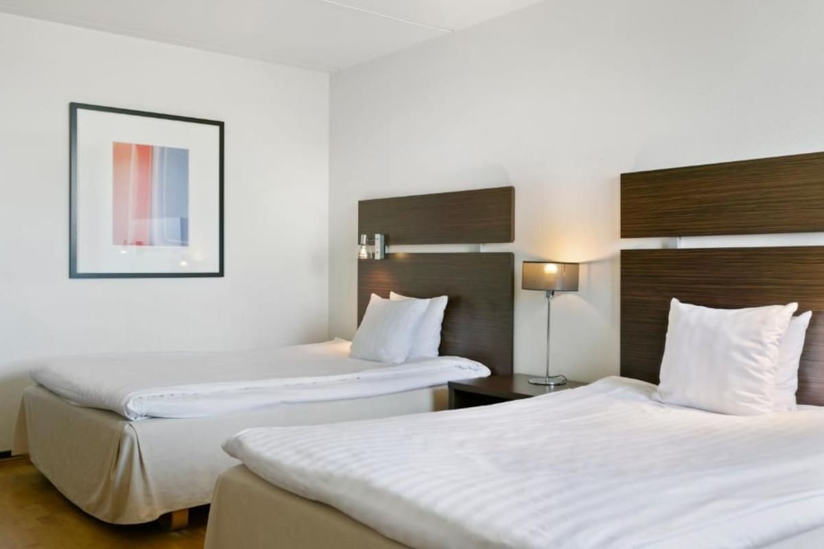 Habitación de hotel con dos camas individuales, una mesita de noche con lámpara y un cuadro en la pared.