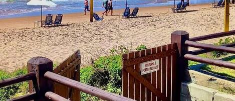 A beach with a wooden staircase leading down to the sand, a sign indicating a closed gate, and beach chairs and umbrellas on the sand.