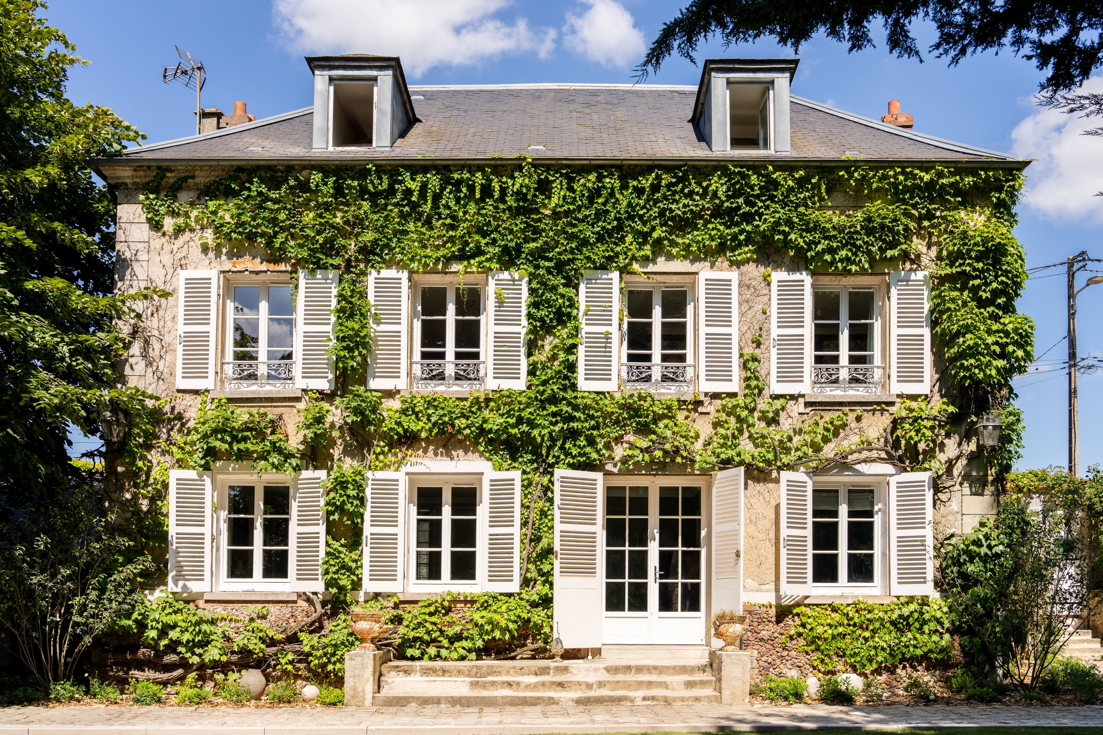 Un bâtiment à deux étages, doté de volets blancs, d’une façade en pierre et d’un jardin à l’avant.