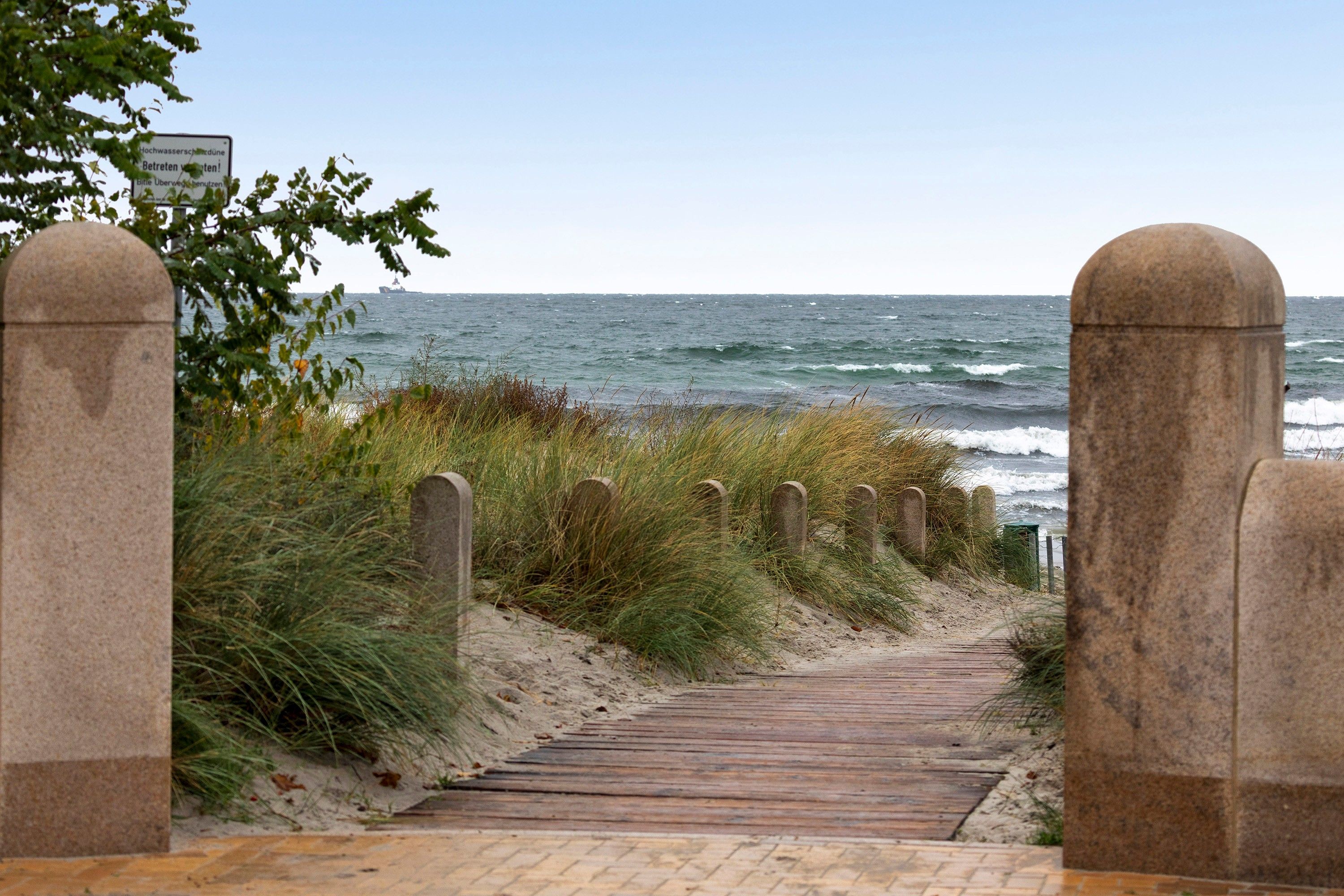 Ein Küstenweg führt zum Meer, gesäumt von Steinpfeilern und einem Schild.