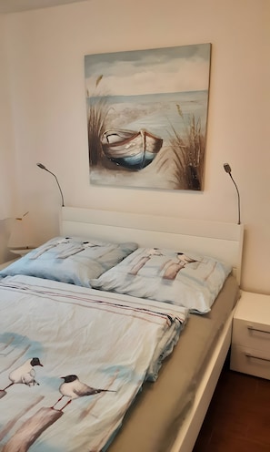 A bed with a white headboard, a painting of a boat on the wall, and a bedside table with a lamp.