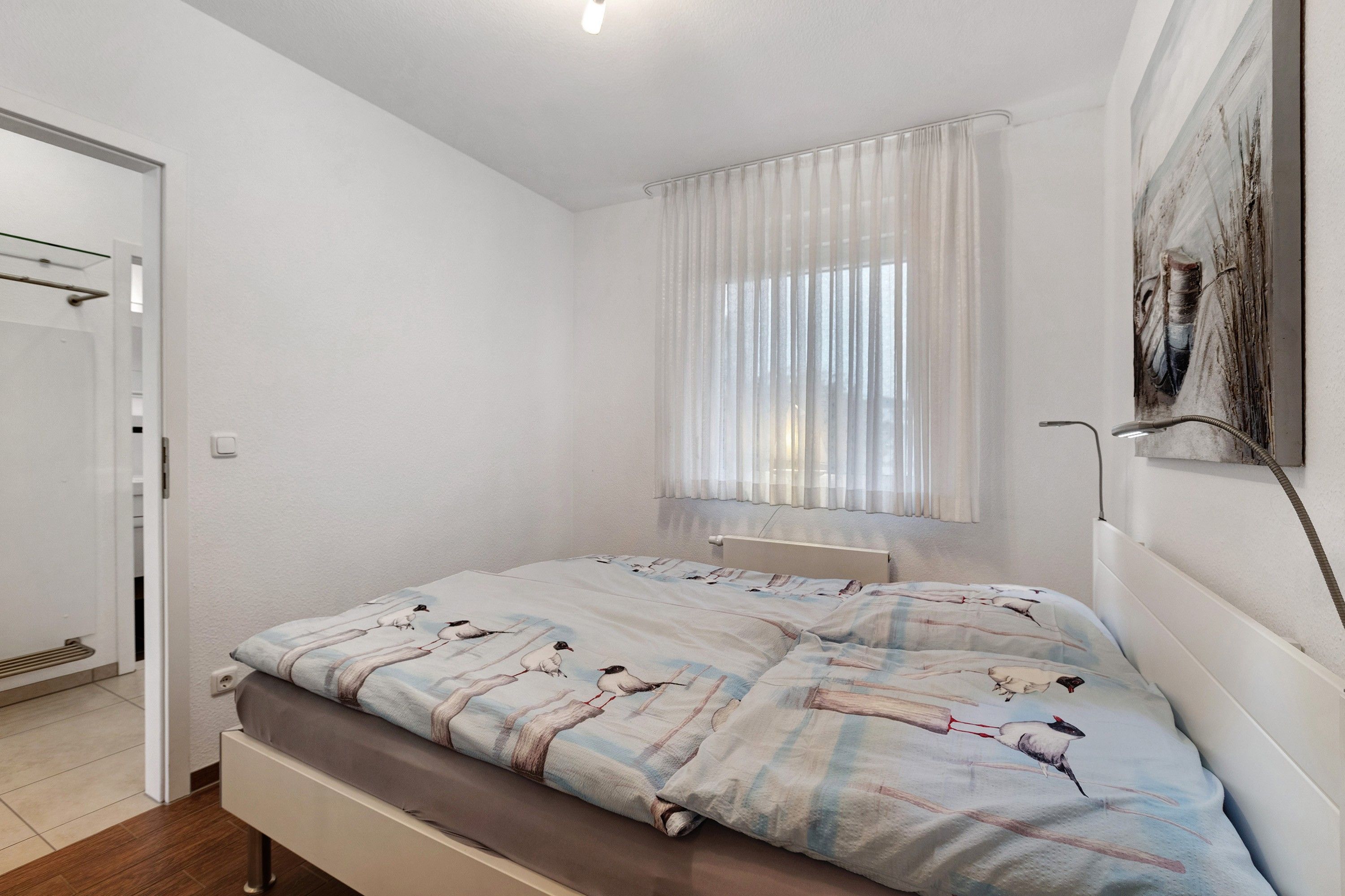 Ein Schlafzimmer mit Bett, einem Fenster mit Vorhängen, einem Heizkörper und einem Bild an der Wand.
