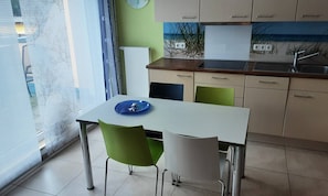 A small dining area with a table and chairs, a window with blinds, and a kitchen area with cabinets and a sink.