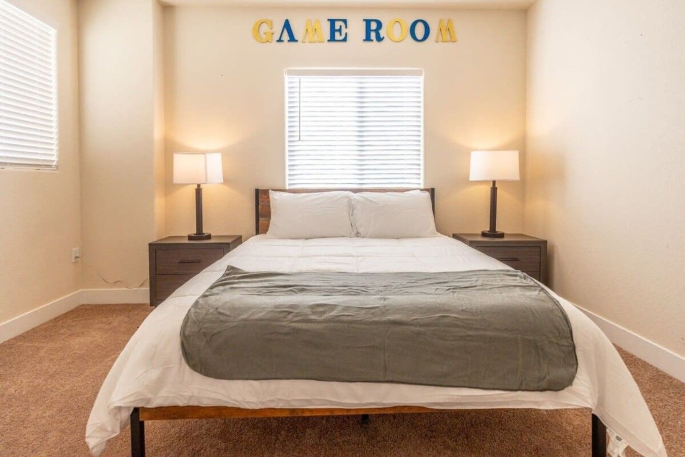 Une chambre à coucher avec un lit, deux tables de chevet avec des lampes, une fenêtre avec des stores et une pancarte au-dessus du lit sur laquelle est écrit « SALLE DE JEUX ».
