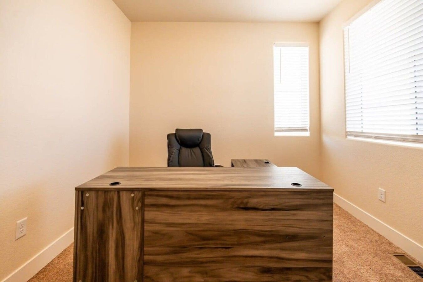 A room with a large wooden desk, a black office chair, and a window with blinds.