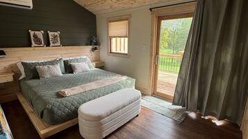 Une chambre avec un plafond en bois, un lit recouvert d’une courtepointe verte matelassée, un pouf blanc, une fenêtre avec des stores et une vue sur la verdure à l’extérieur.