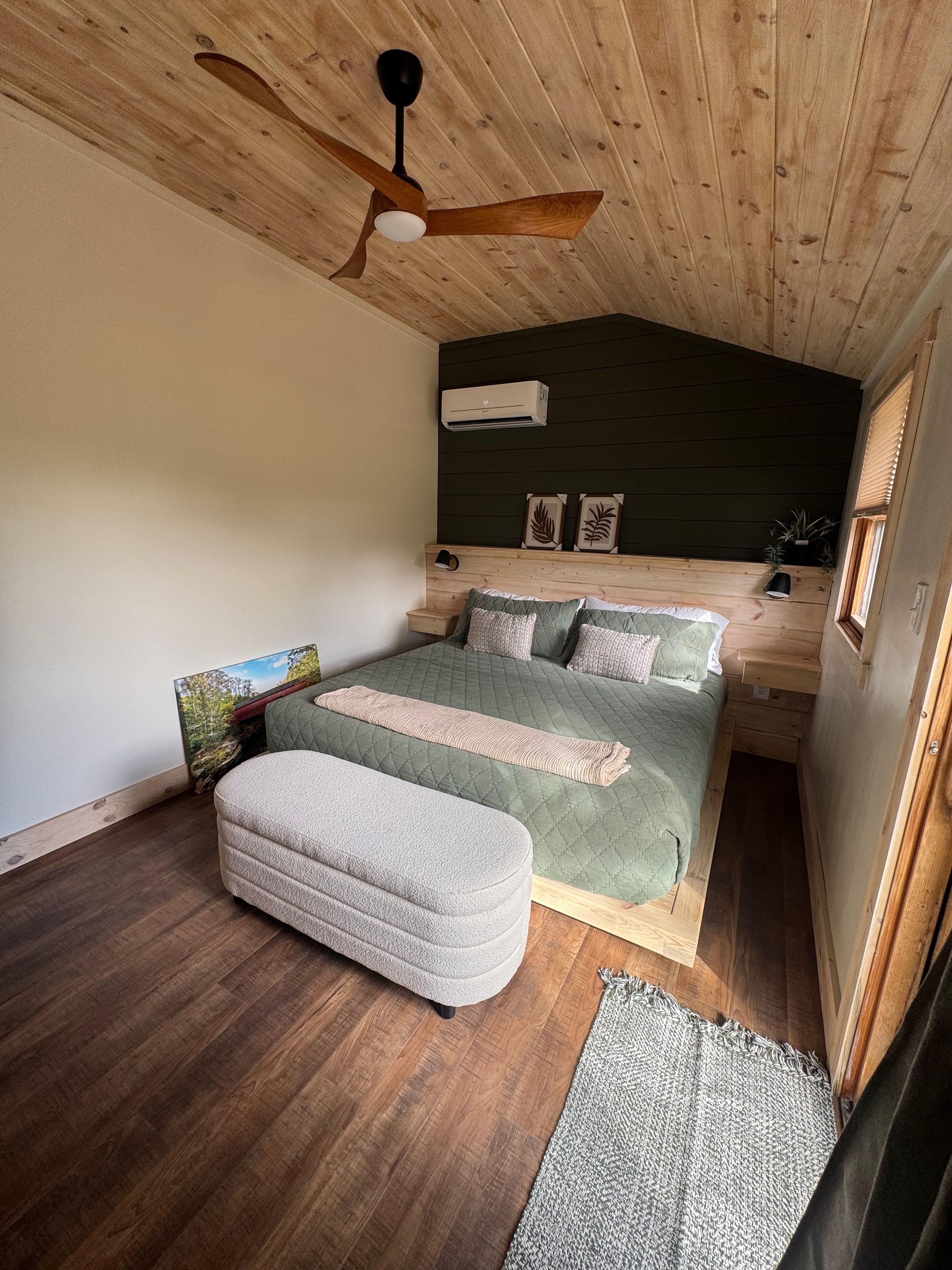 A bedroom with a wooden ceiling, a bed with green bedding, a white ottoman, and a ceiling fan.
