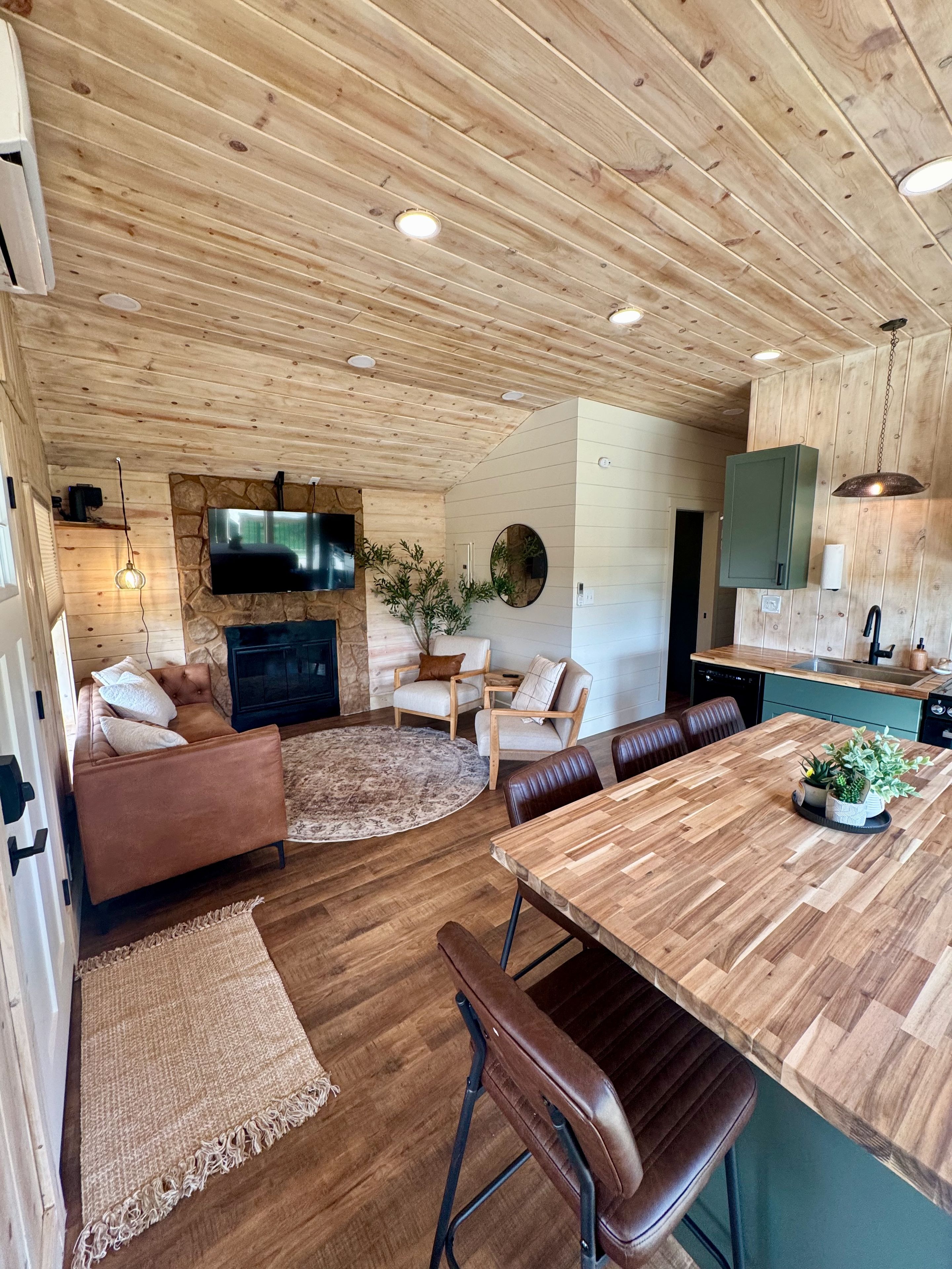 A modern interior with a wooden ceiling, a flat-screen TV mounted on a stone wall, a dining area with a wooden table and chairs, and a kitchen with a green cabinet.