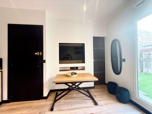 A modern kitchen with a black door, a wooden table with a bowl, a mounted TV, and a large window with a view of the outdoors. - B087 Appartement cosy et moderne – Terrasse-Centre-ville (Cannes)