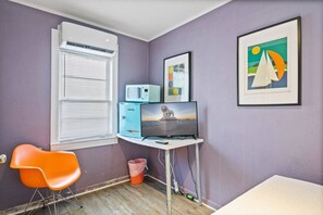 Una habitación con escritorio, silla, ventana con persianas, un cesto de basura y cuadros en la pared.