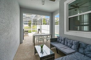 A covered outdoor seating area with a table and chairs, a cushioned bench, and a view of a swimming pool. - 4BR Champions Gate Villa | Oasis Club Amenitie’s (Kissimmee)