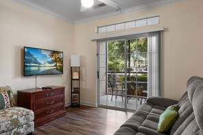 A living room with a sofa, a TV mounted on the wall, a wooden cabinet, and a sliding glass door leading to an outdoor area with chairs. - 2BR Tuscana Condo | Relax | Pool | Amenities (Kissimmee)