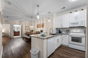 A modern kitchen with white cabinetry, a central island, and a dining area with wooden chairs. - 2BR Tuscana Condo | Relax | Pool | Amenities (Kissimmee)