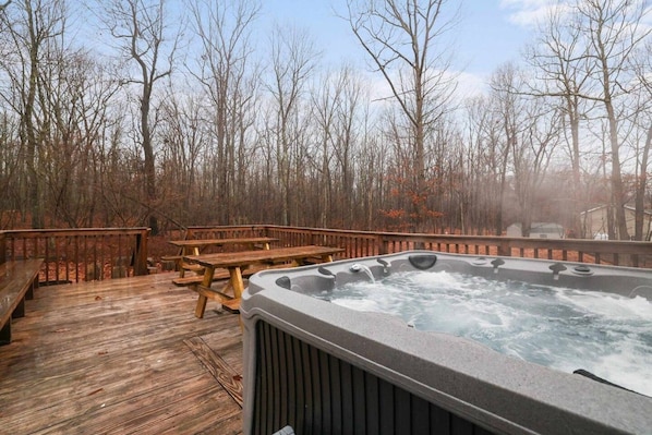 A wooden deck with a hot tub and picnic tables, surrounded by leafless trees.