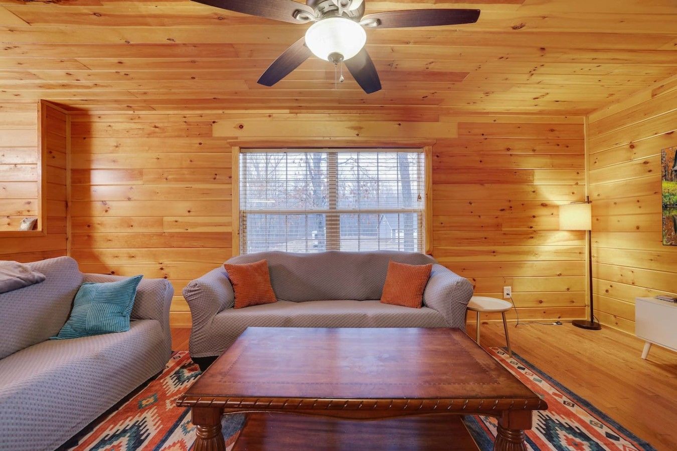 Ein gemütliches Zimmer mit Holzboden, Deckenventilator, einer Couch, einem Couchtisch und einem Fenster mit Jalousien.