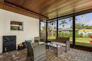 A covered patio area with wicker furniture, a glass table, and a view of a backyard with palm trees. - Fort Myers Waterfront Retreat | 4BR Home W/Views (Fort Myers)