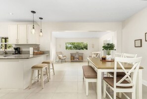 A modern kitchen with white cabinetry, a central island with stools, and a dining area with white chairs. - Fort Myers Waterfront Retreat | 4BR Home W/Views (Fort Myers)