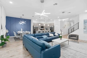 A modern living room with a blue sectional sofa, a dining area, and a kitchen in the background. - Super Ritzy 9BR | Ultimate Disney Retreat (Orlando)