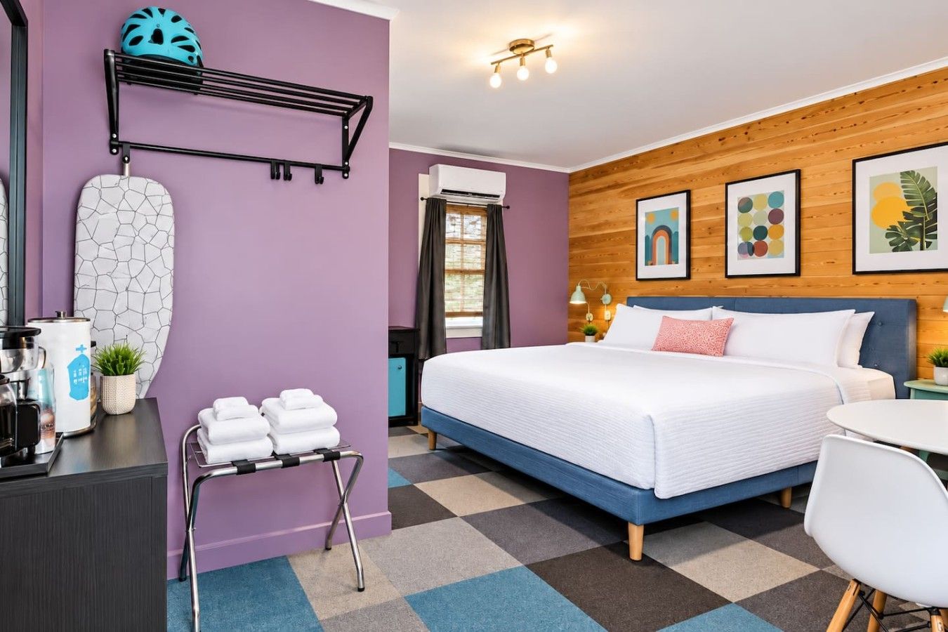 A hotel room with a bed, a desk, a chair, a wall-mounted rack, and a window.