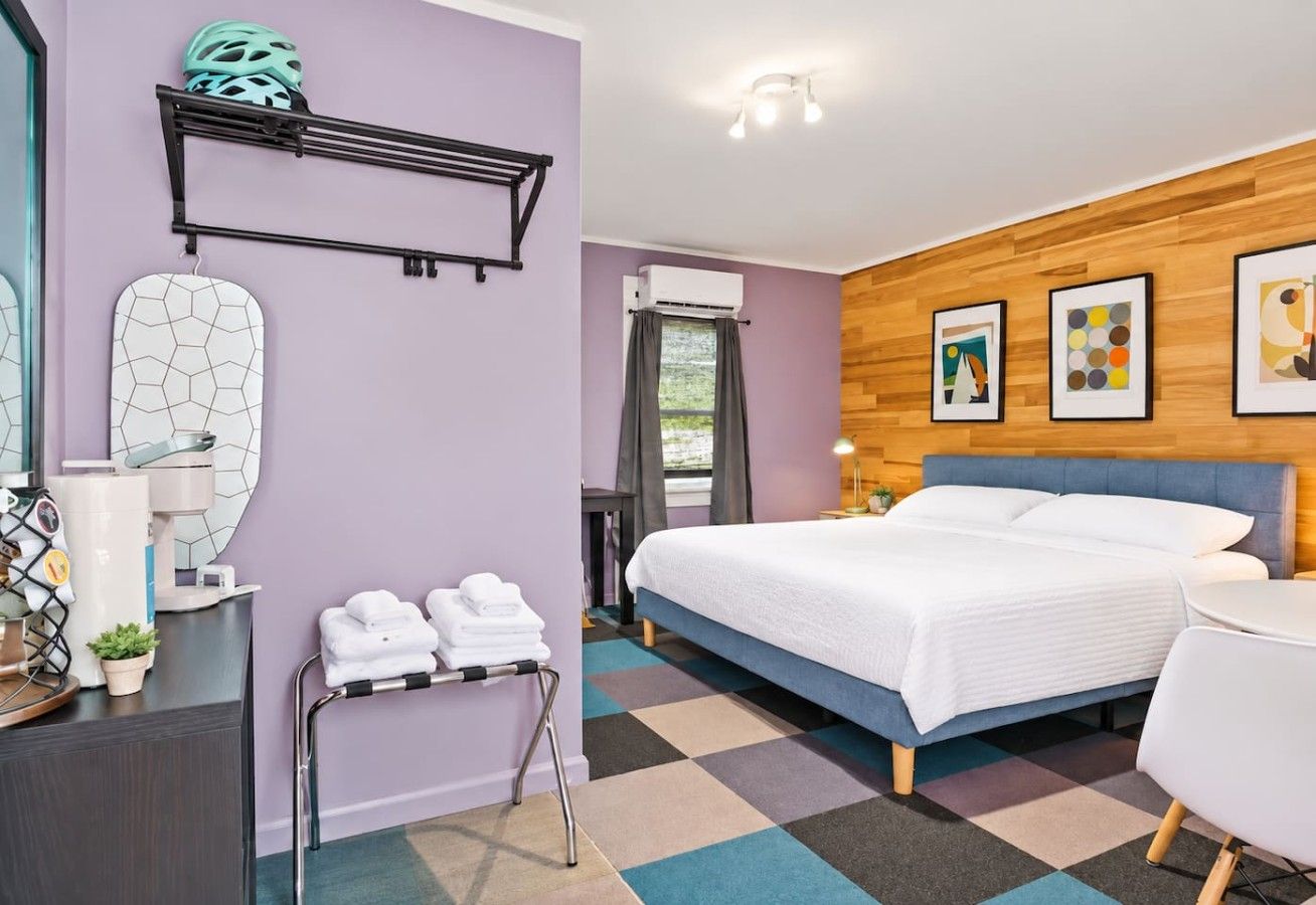 A hotel room with a bed, a desk, a chair, a shelf with towels, and artwork on the wall.