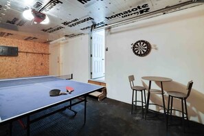 Una mesa de ping pong con dos palas, un dardo y dos sillas en una habitación con pared de madera y puerta blanca.